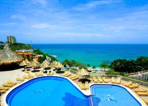 A resort pool with two sections, surrounded by lounge chairs and thatched umbrellas, overlooking a beach and ocean. - Hippocampus Vacation Club (Pampatar)