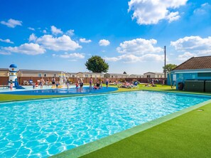 A swimming pool with a water play area, surrounded by a fence and a building with a tiled roof. - Golden Dune - Hayling Island (Hayling Island)