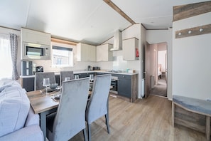 A compact kitchen with modern appliances, a dining area with a table and chairs, and a visible bedroom in the background. - Wave Whisperers - Hayling Island (Hayling Island)