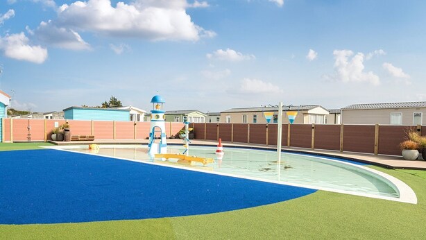 A swimming pool with a blue and green surface, surrounded by a wooden fence and houses in the background. - The Lookout - Hayling Island (Hayling Island)