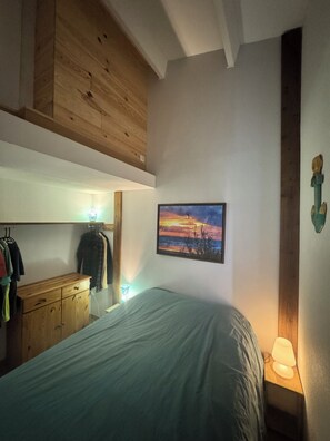 A bedroom with a bed, a wooden dresser, a wall-mounted lamp, and a painting.