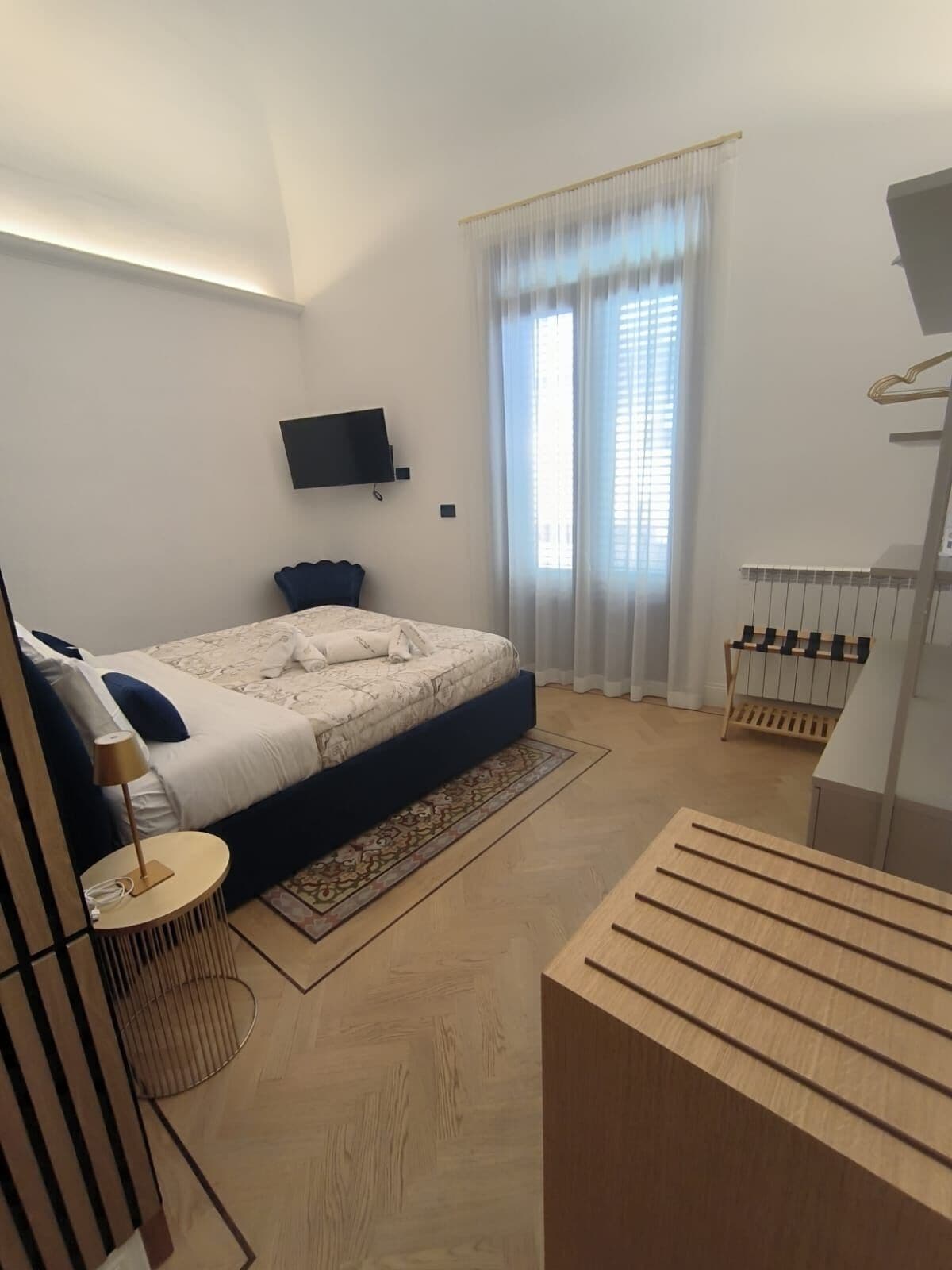 A modern bedroom with a bed, a TV mounted on the wall, a window with sheer curtains, and a wooden bench.