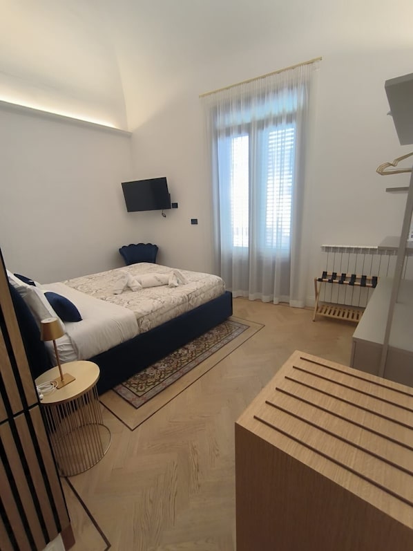 A modern bedroom with a bed, a TV mounted on the wall, a window with sheer curtains, and a wooden bench.