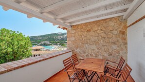 Una terraza con muro de piedra, mesa de madera con sillas y vista a una playa y montañas.