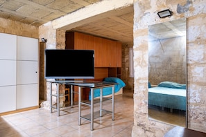 Une piĂšce avec un tĂ©lĂ©viseur posĂ© sur un support mĂ©tallique, un lit recouvert dâune couverture bleue et un miroir qui reflĂšte le lit.