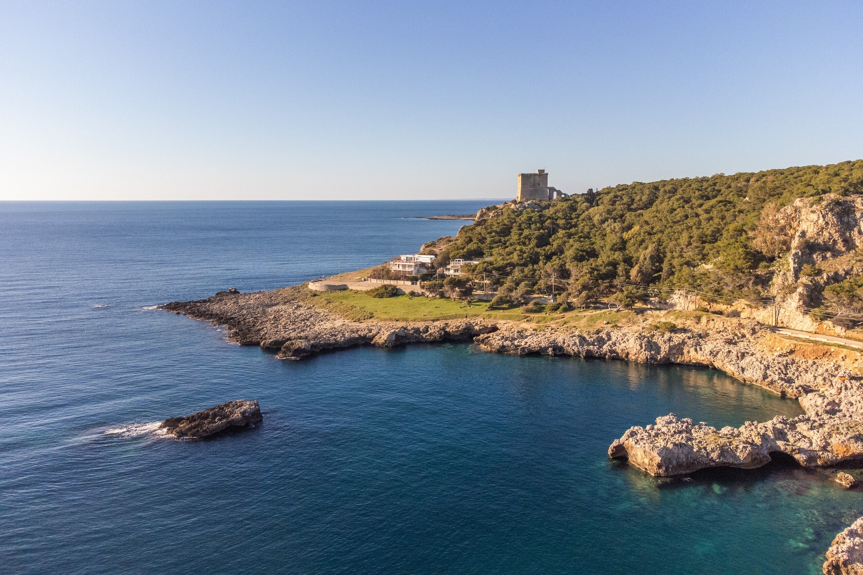 Un paysage côtier avec un château perché sur une colline, une côte rocheuse et une mer d’un bleu limpide.
