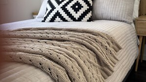 Een net opgemaakt bed met een gekleurd kussen, een gebreide plaid en een houten hoofdbord.