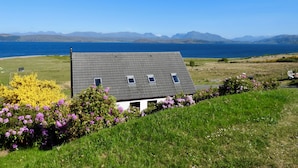 Une maison avec un toit en pente, une cour clôturée et une vue sur la mer et les montagnes en arrière-plan.