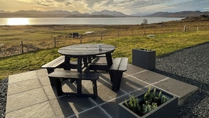 Une table de pique-nique avec des bancs, un petit jardinière avec des plantes, et une vue sur un lac avec des montagnes en arrière-plan.