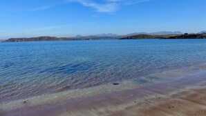 Un lac paisible aux eaux d’un bleu limpide, une plage de sable et, au loin, des montagnes sous un ciel dégagé.