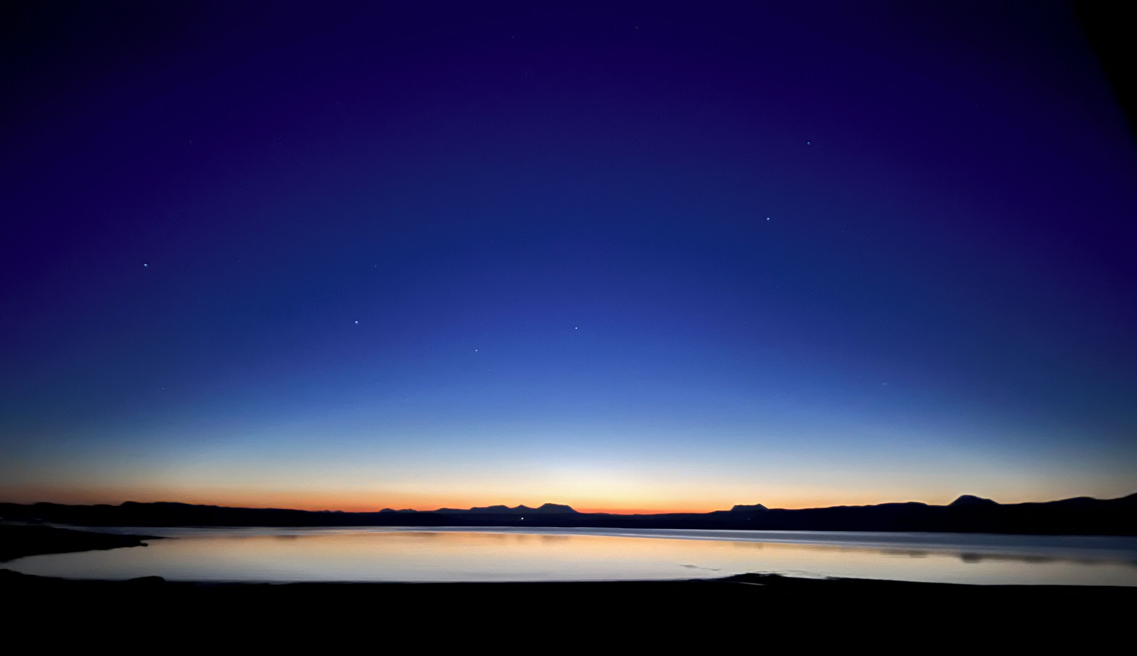 Paisatge nocturn amb un llac tranquil que reflecteix el cel, una serralada a la llunyanàcia i unes quantes estrelles visibles.