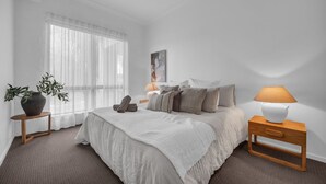 Ein Schlafzimmer mit einem großen Bett, einem Nachttisch mit Lampe, einem kleinen Tisch mit Pflanze und einem Fenster mit durchsichtigen Vorhängen.