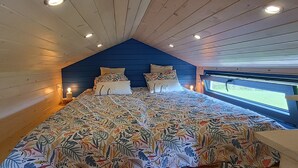 A bedroom with a sloped ceiling, a large bed with patterned bedding, two bedside lamps, and a window with a view of greenery.
