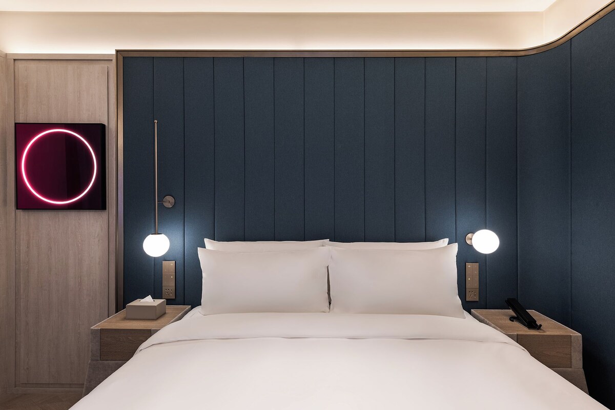 Une chambre d’hôtel moderne avec une tête de lit bleu foncé, deux tables de chevet avec des lampes et un lit recouvert de draps blancs.