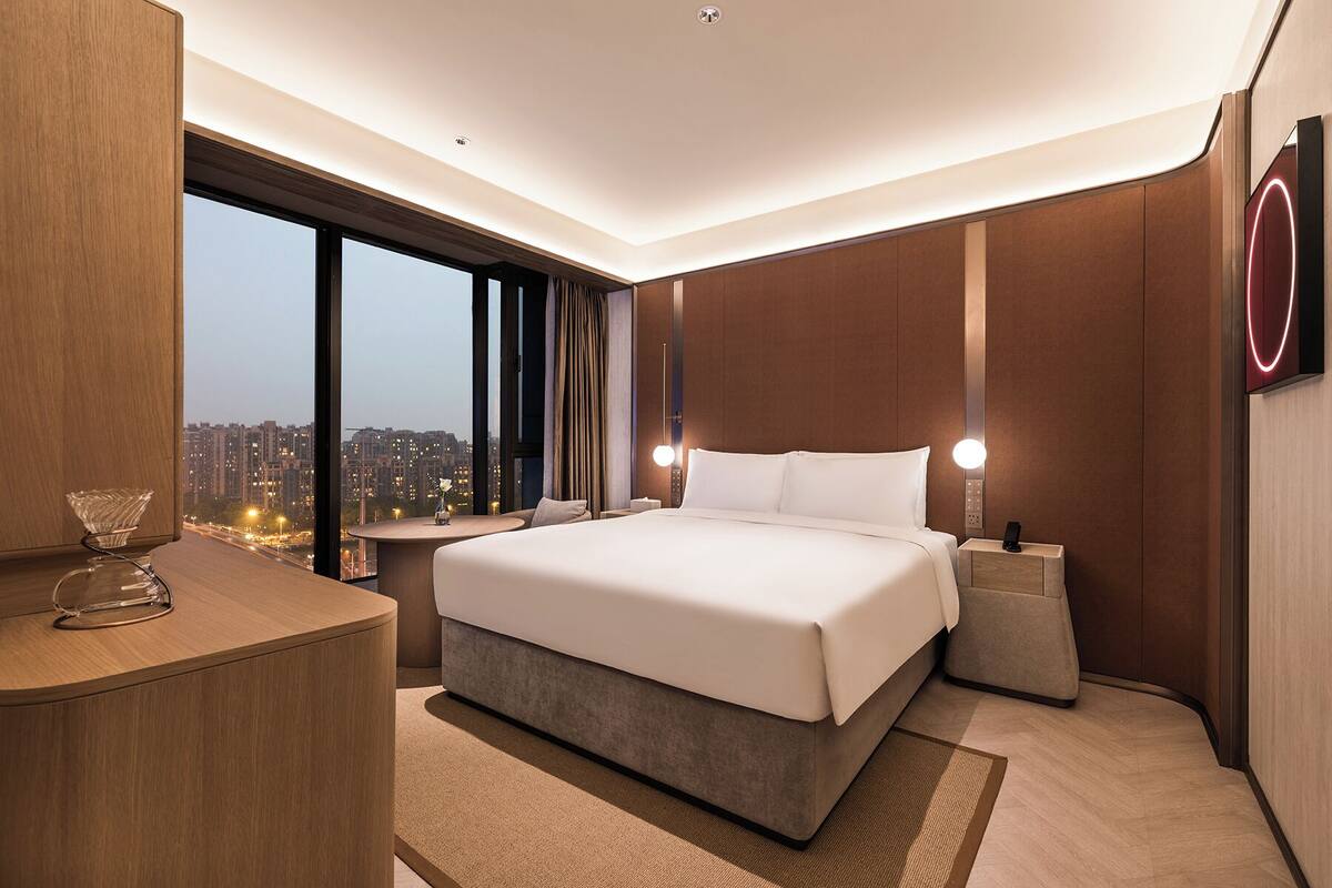 A modern hotel room with a large bed, a desk, and a view of the cityscape.
