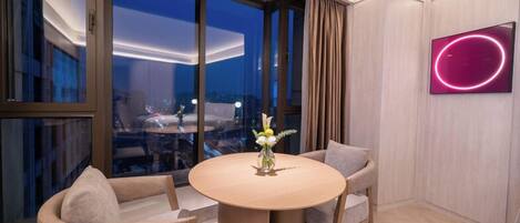 A modern hotel room with a round table, two chairs, a vase with flowers, a large window with curtains, and a neon sign on the wall.