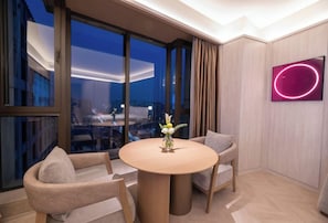 A modern hotel room with a round table, two chairs, a vase with flowers, a large window with curtains, and a neon sign on the wall.