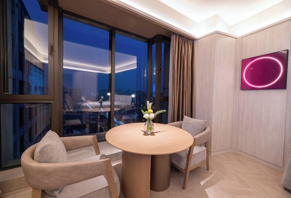 Una habitación de hotel moderna con una mesa redonda, dos sillas, un florero con flores, un ventanal con cortinas y un letrero de neón en la pared.