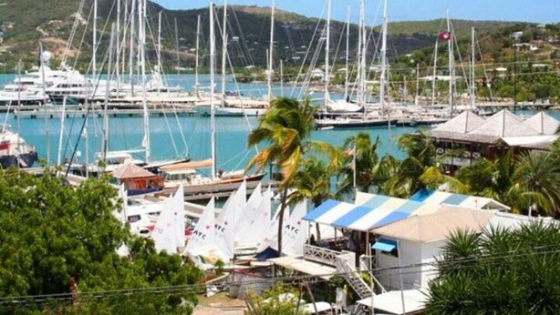 A marina with multiple sailboats docked, surrounded by lush greenery and hills in the background.
