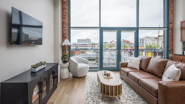 A modern living room with a large window, a brown leather sofa, a wooden coffee table, and a black entertainment unit. - Cozy, Modern Bay Views for Six! (Sandusky)