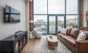 A modern living room with a large window, a brown leather sofa, a wooden coffee table, and a black entertainment unit. - Cozy, Modern Bay Views for Six! (Sandusky)