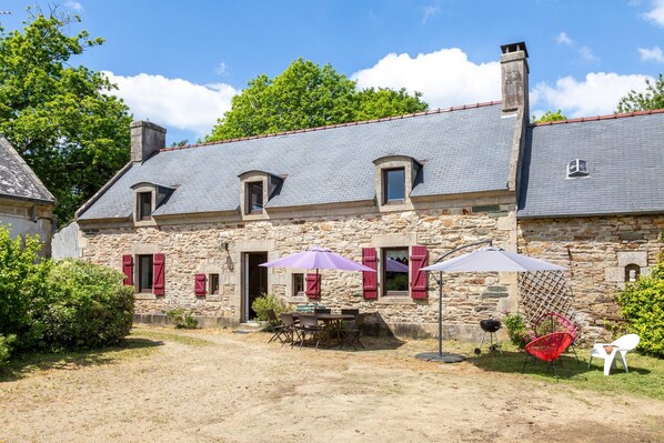 A stone building with a tiled roof, a patio with a table and chairs, and a barbecue grill.