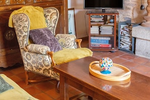 A living room with a patterned armchair, a wooden coffee table with a decorative globe, a bookshelf with books, and a television. - Holiday Home 'Charme Armorine' with Private Garden (Plonéour-Lanvern)