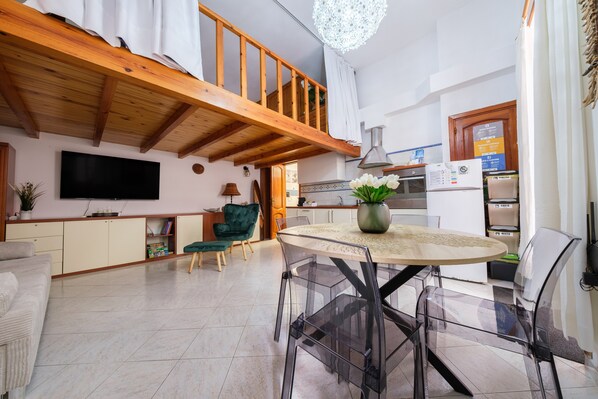 A spacious living area with a high ceiling, a wooden staircase, a kitchen with modern appliances, and a dining area with transparent chairs.