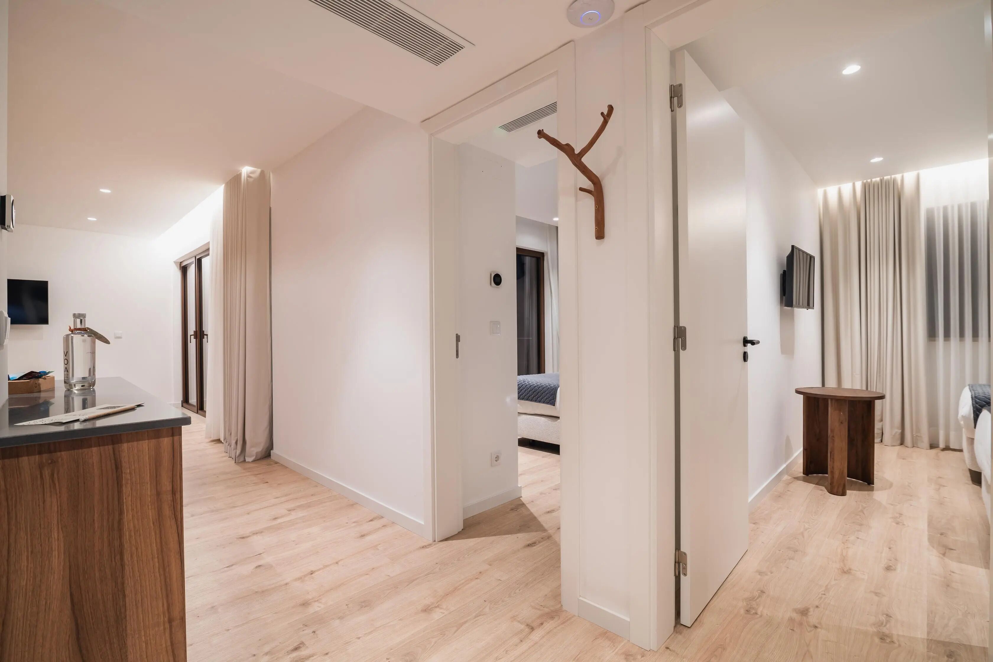 A modern hotel room with a wooden floor, a small wooden side table, a bed with a blue blanket, a TV mounted on the wall, and a door leading to another room.