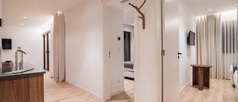 Una habitación de hotel moderna con piso de madera, una mesita de madera pequeña, una cama con manta azul, un televisor montado en la pared y una puerta que da a otra habitación.