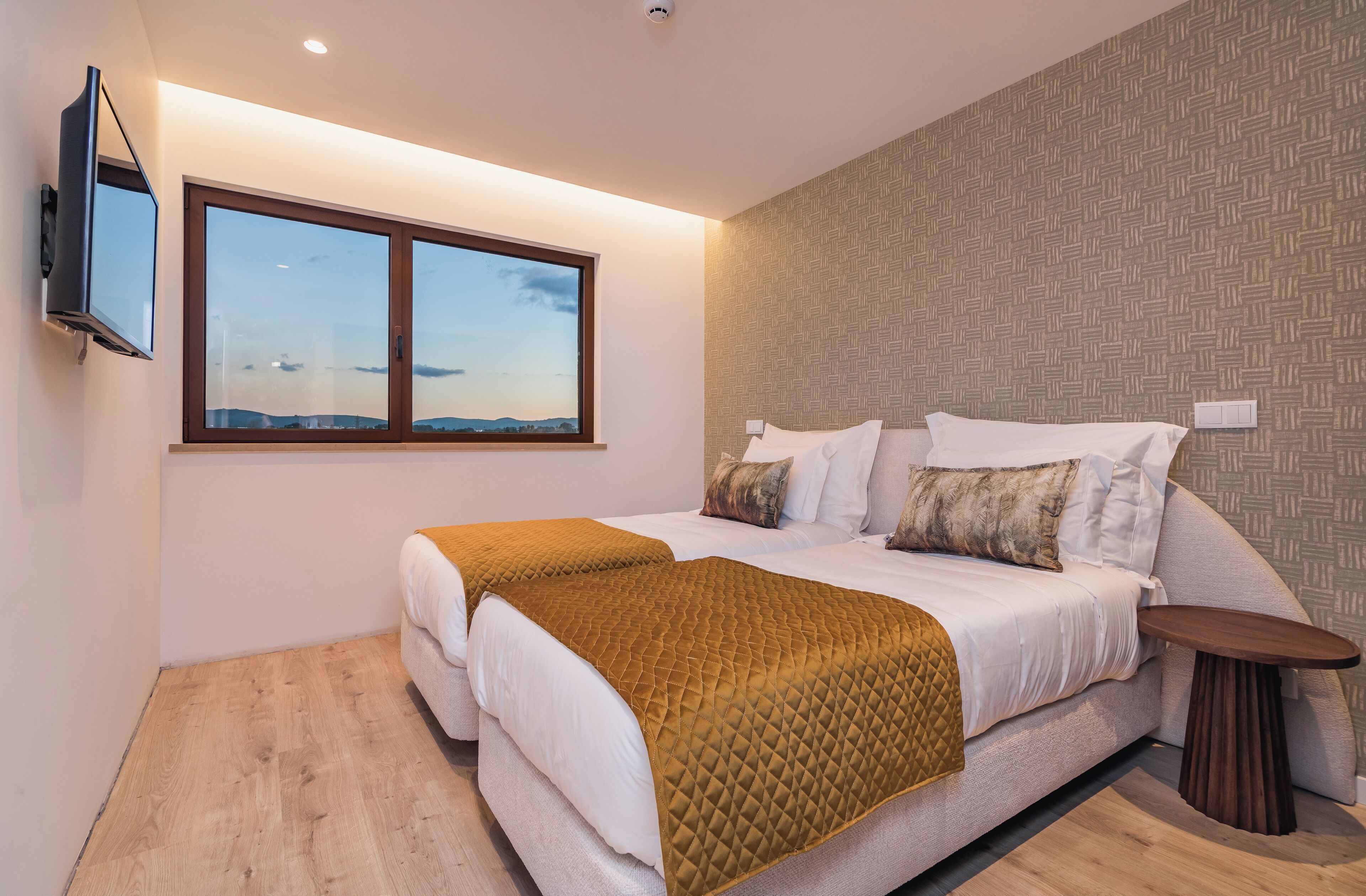 A modern bedroom with two beds, a large window, a flat-screen TV, and a small table.