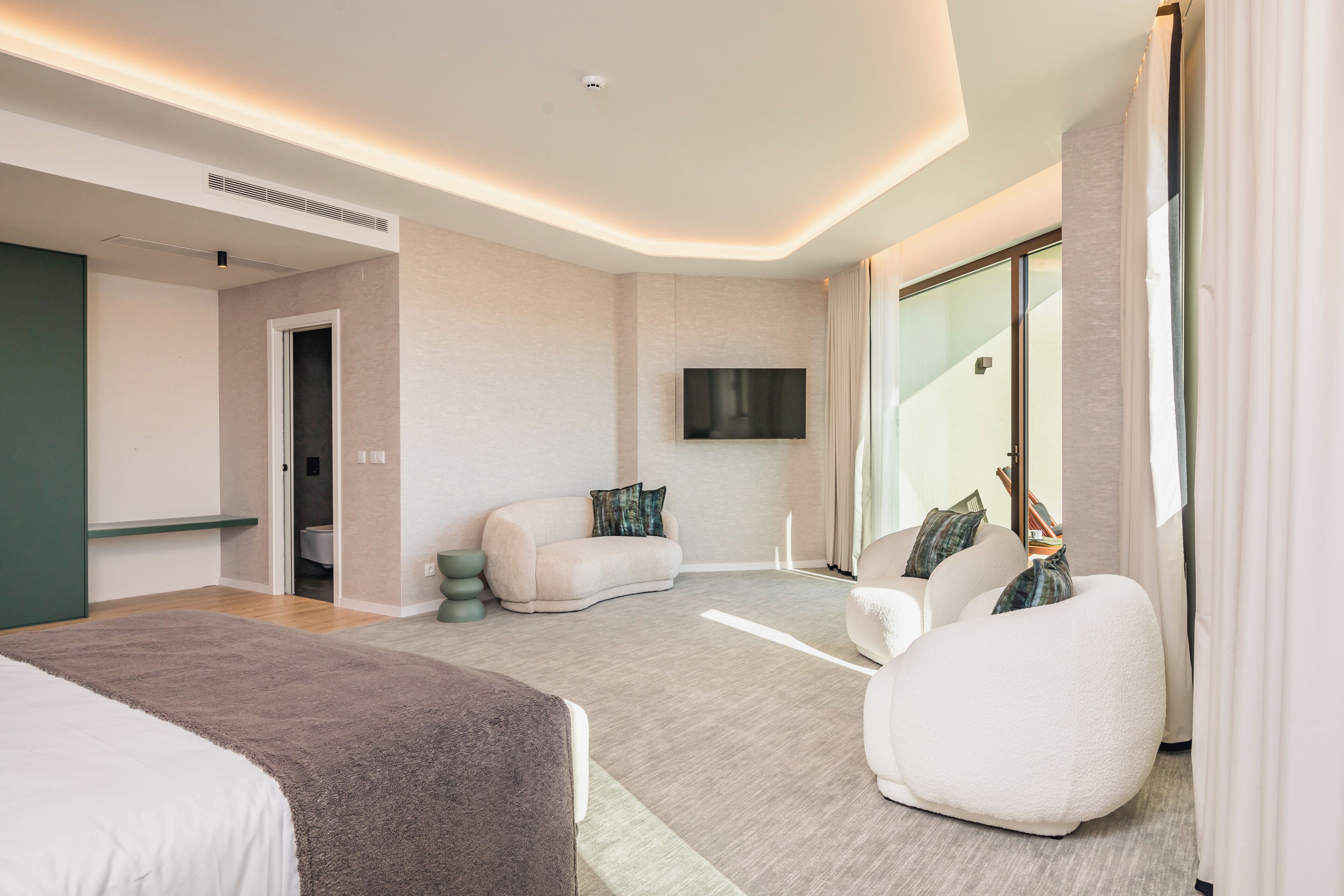 A modern hotel room with a large bed, a seating area with a sofa and armchair, a flat-screen TV mounted on the wall, and a large window with curtains.