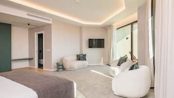 A modern hotel room with a large bed, a seating area with a sofa and armchair, a flat-screen TV mounted on the wall, and a large window with curtains.