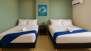 A hotel room with two beds, a small table, a nightstand, a framed picture of an island, and an air conditioning unit.