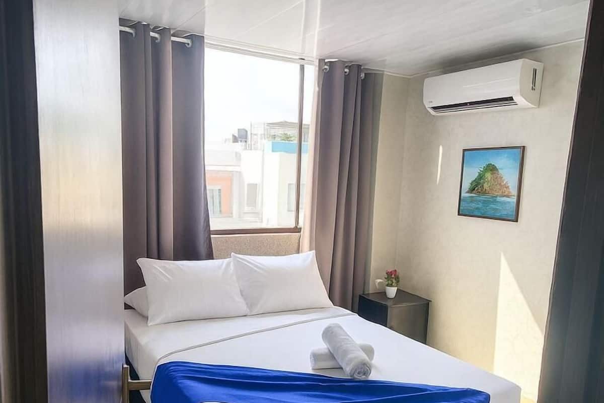 A hotel room with a bed, curtains, a nightstand, an air conditioning unit, and a painting on the wall.