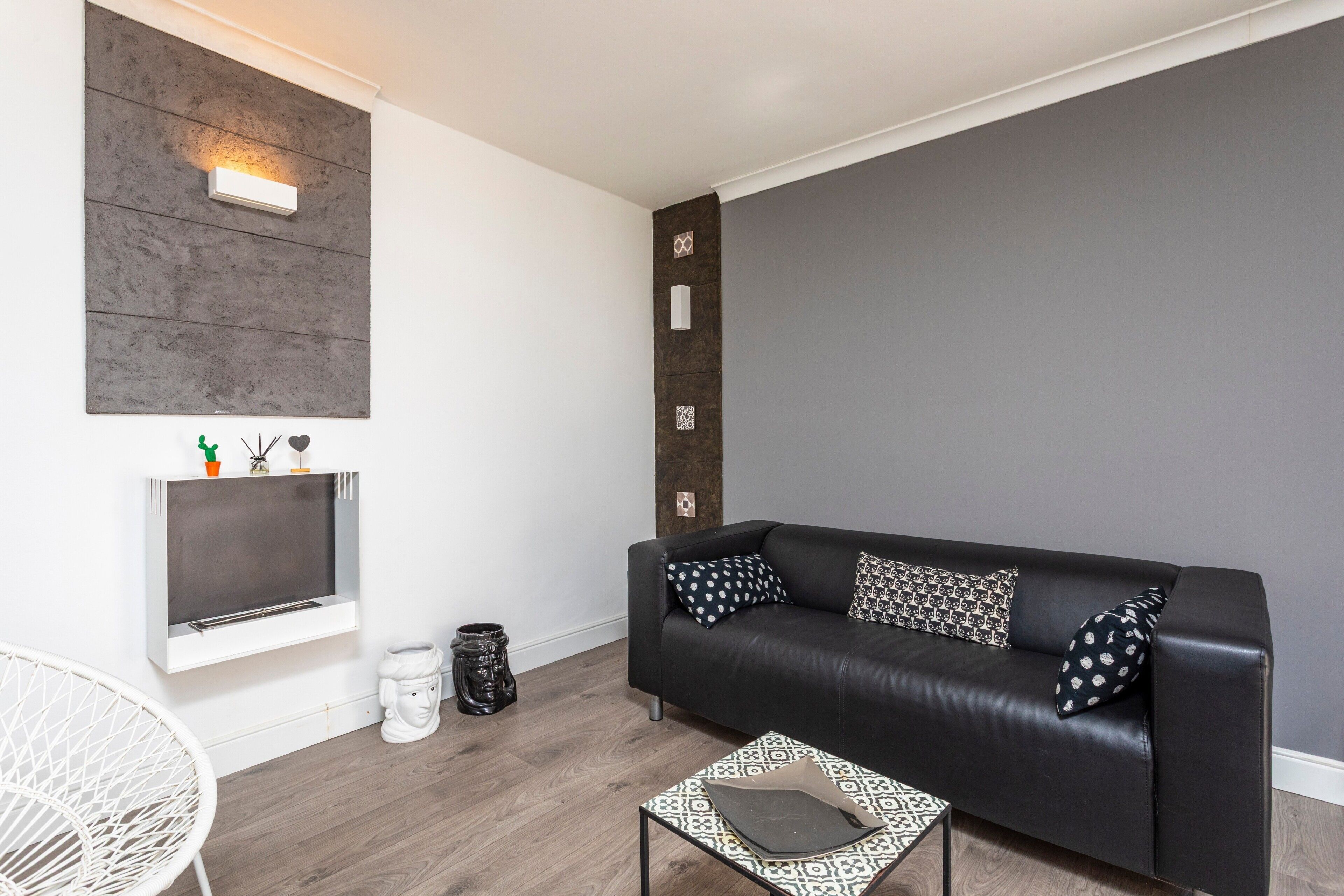 A modern living room with a black leather sofa, patterned cushions, a white chair, a small table, and a wall-mounted shelf with decorative items.