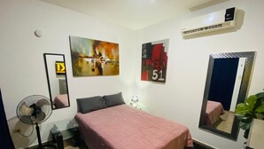 Une chambre à coucher comprenant un lit, un ventilateur sur pied, un miroir et deux œuvres d’art accrochées au mur.
