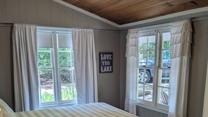 A bedroom with a bed, two windows with curtains, a sign saying "LOVE THE LAKE", and a view of the outdoors.