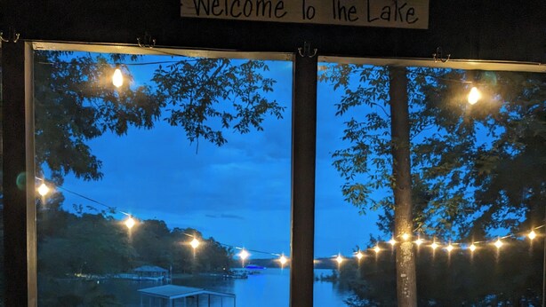 Uitzicht door een raam met een 'Welcome To The Lake' bord, een houten terras en lichtslingers.