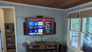 Een woonkamer met een wandgemonteerde flatscreen televisie waarop verschillende zenders draaien, een houten entertainmentkast met opbergruimte, en een raam met uitzicht op de omgeving.