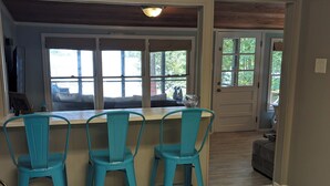 Een eetgedeelte met een houten tafel en turquoise stoelen, een keuken met grote ramen en een deur naar een andere kamer.