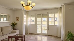 A spacious living room with a sofa, a glass-top coffee table, a chandelier, and large windows with curtains.