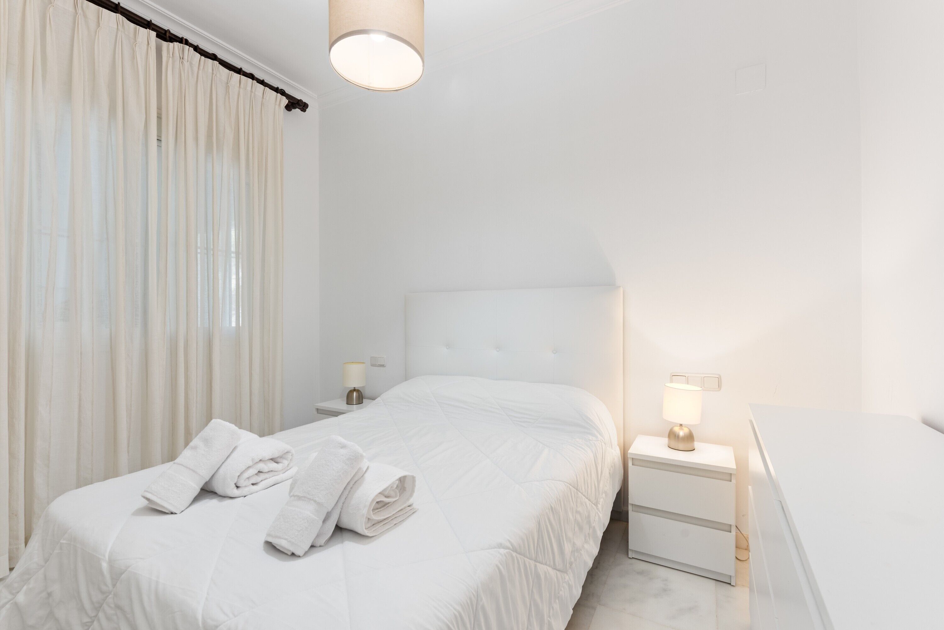 Una habitación de hotel con cama, mesita de noche con lámpara y ventana con cortinas.