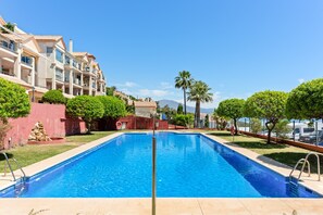 A swimming pool with a clear blue water, surrounded by a well-maintained lawn, palm trees, and a multi-story building with balconies. - Apartment 'Panoramic Sea Views' with Sea View, Wi-Fi and Air Conditioning (Manilva)