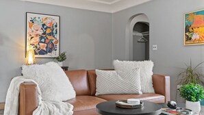 A modern living room with a brown leather sofa, white fluffy throw, and a round coffee table. There are two framed artworks on the walls, a potted plant, and a hallway visible through an archway.