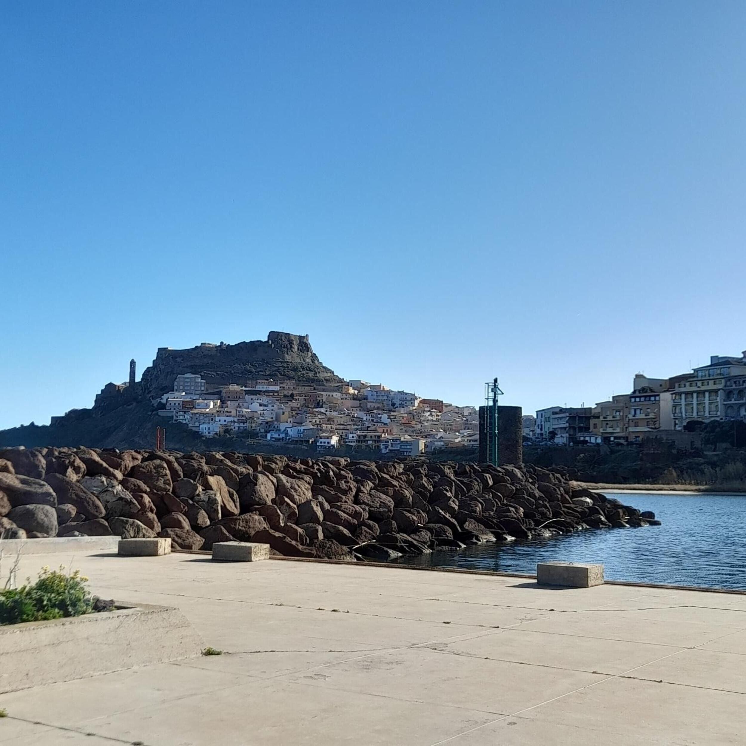 A coastal cityscape with a prominent rocky hill, a clear sky, and a calm sea.