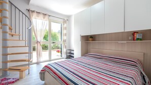 A bedroom with a bed, a wooden headboard, a window with curtains, a staircase, and a shelf with books. - Vacation Home 'Mare Nostrum' with Mountain View, Private Terrace, and Air Conditioning (Castelsardo)