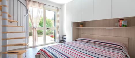 A bedroom with a bed, a wooden headboard, a window with curtains, a staircase, and a shelf with books.