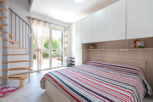 A bedroom with a bed, a wooden headboard, a window with curtains, a staircase, and a shelf with books.