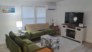 A living room with a green sofa, a small table with a lamp, a TV on a wooden cabinet, and a painting on the wall. - Clambassador Cottage (Cedar Key)